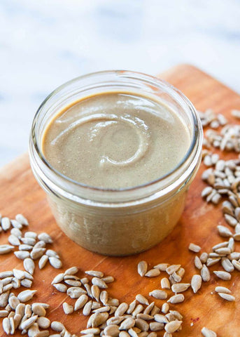 A glass jar of sunflower seed butter resting on a wooden cutting board surrounded by sunflower seeds